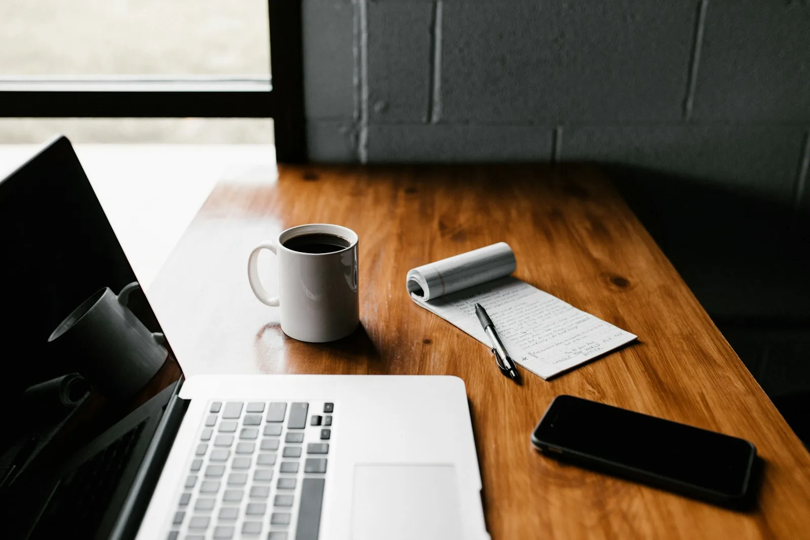 Image of an office desk with laptop, phone, pen, a filled coffee mug and a notepad